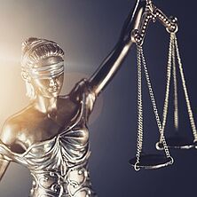 Justitia mit gehobenem Arm im Gegenlicht, in der Hand hält sie eine Wage